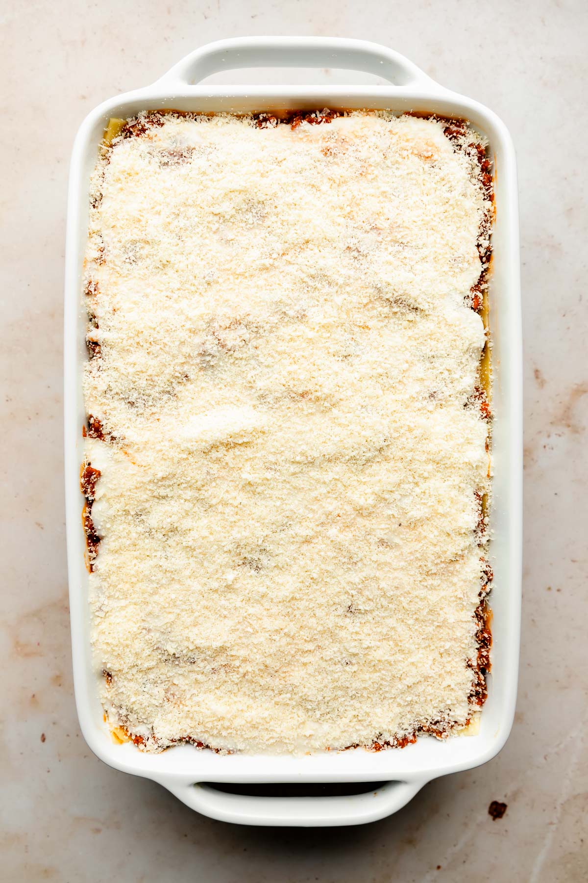 An overhead shot of an assembled, uncooked lasagna in a white baking dish upon an off-white surface.