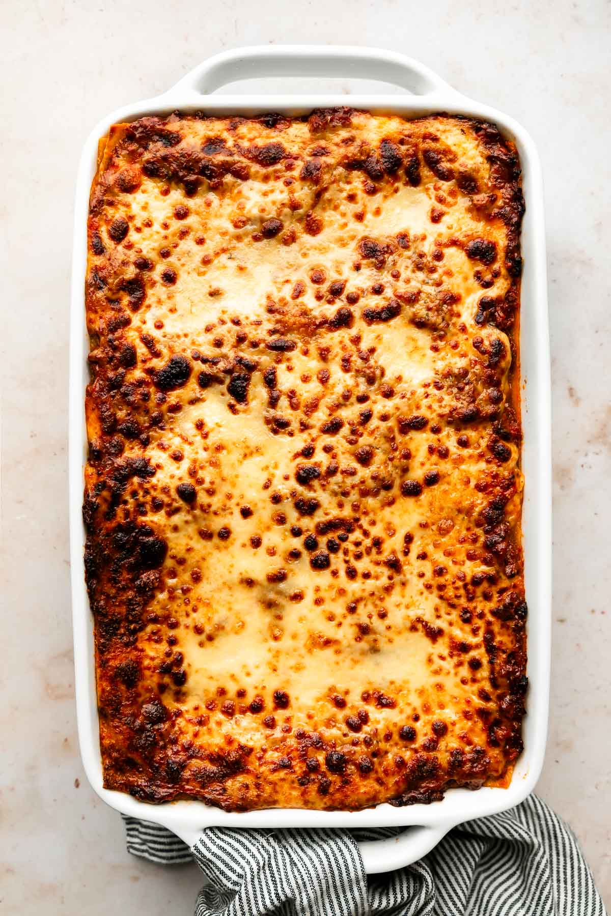 An overhead shot of a cooked lasagna in a white casserole dish on top of a blue and white striped cloth on an off-white surface.