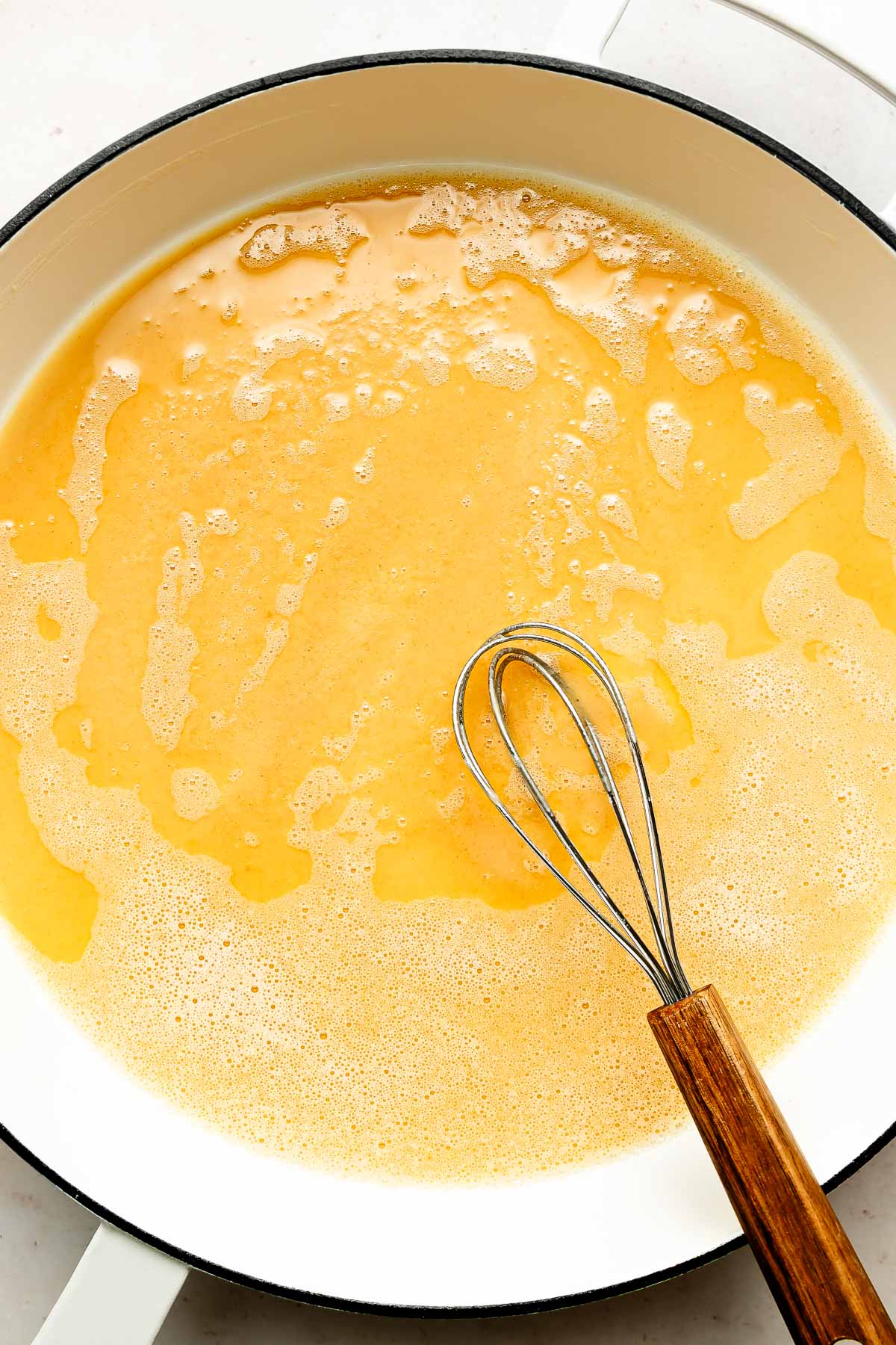 An overhead shot of melted butter with a whisk in a large white skillet atop an off-white surface.