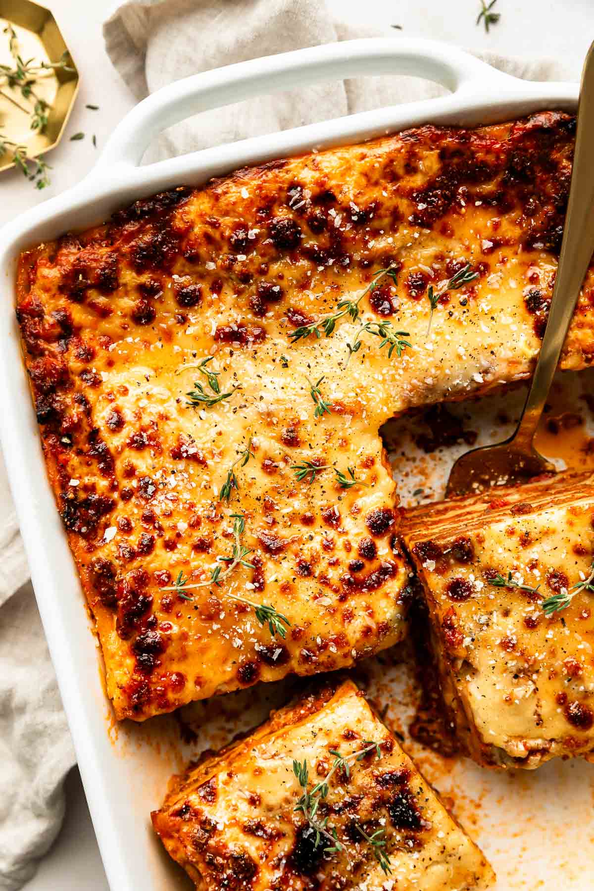 An overhead shot of a cooked lasagna in a white casserole dish atop an off-white surface. Several slices have been cut from the lasagna, including two visible slices.