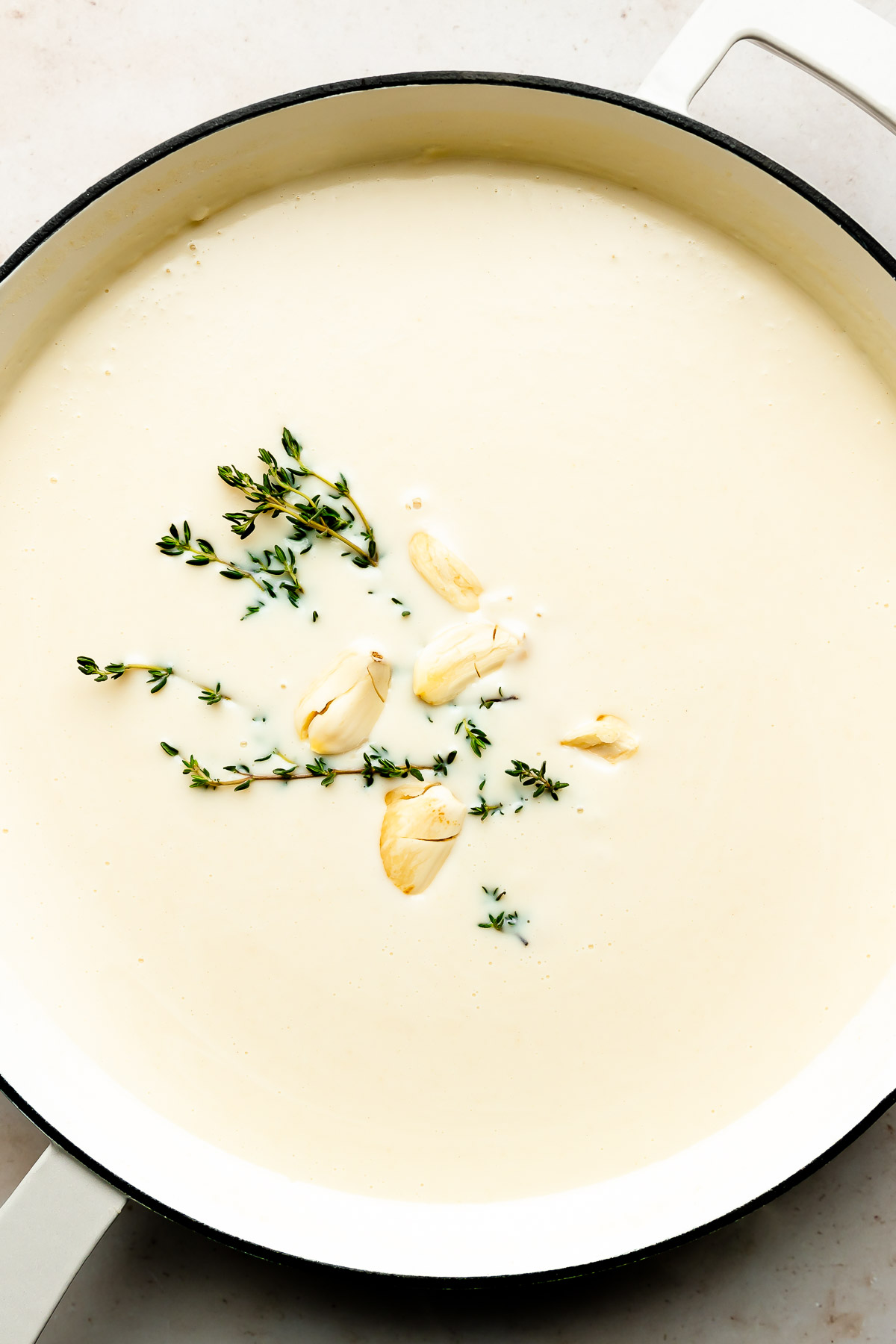 An overhead shot of bechamel sauce with thyme and garlic cloves in a white skillet atop an off-white surface.