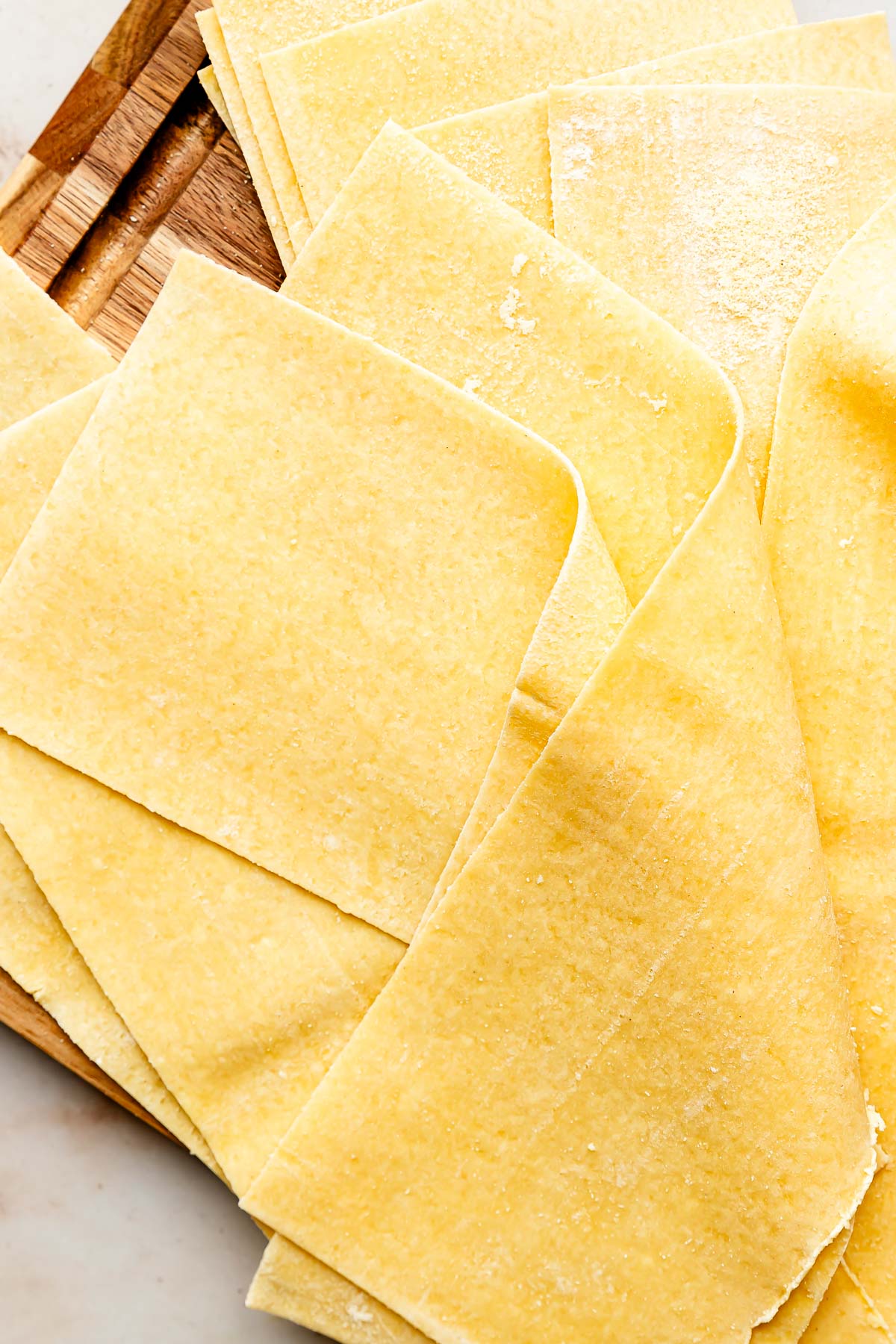An overhead shot of fresh lasagna noodles on a wooden cutting board atop an off-white surface.