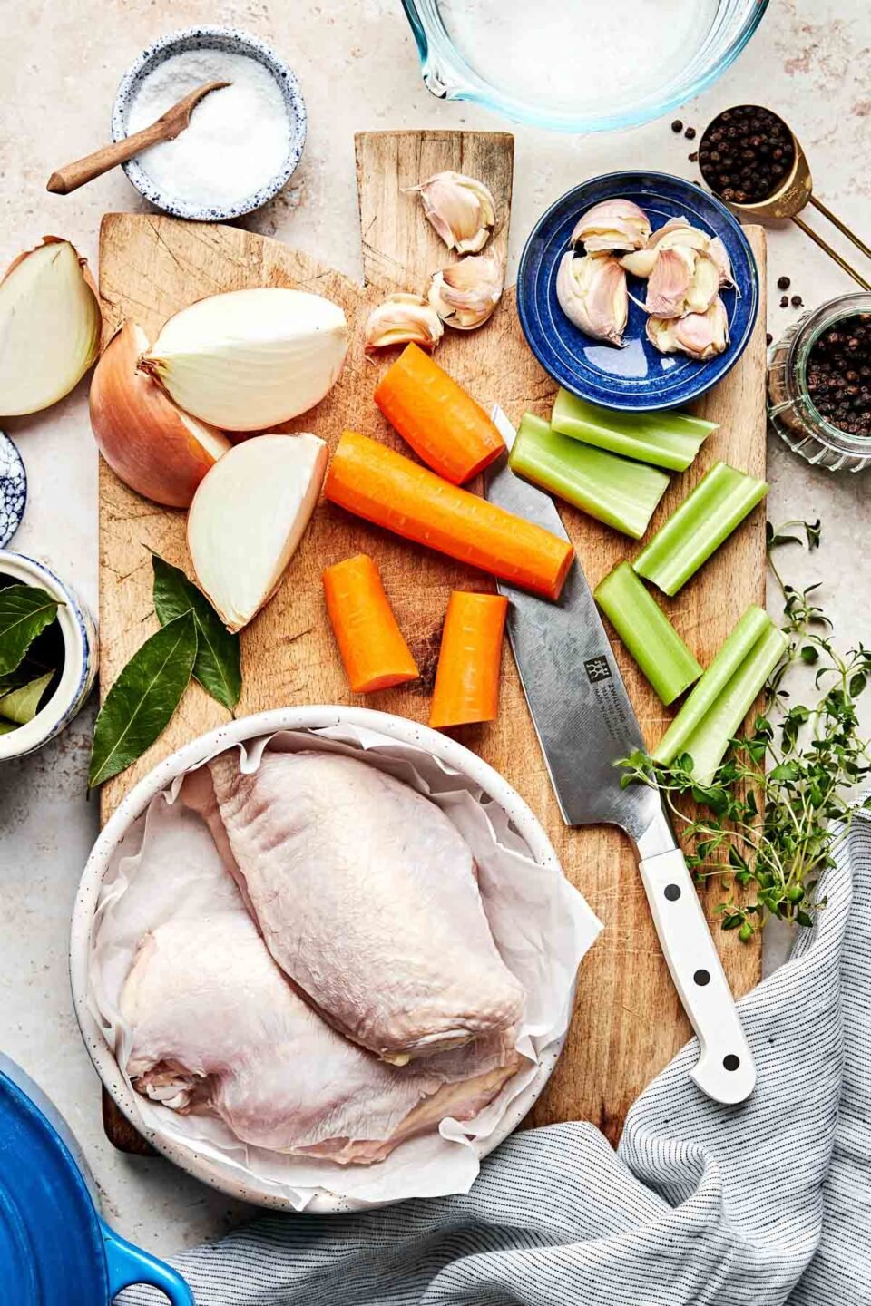 Ingredients are displayed on a wooden board and a textured off-white surface: raw chicken, large pieces of onions, carrots, and celery, garlic, fresh herbs, salt and pepper.