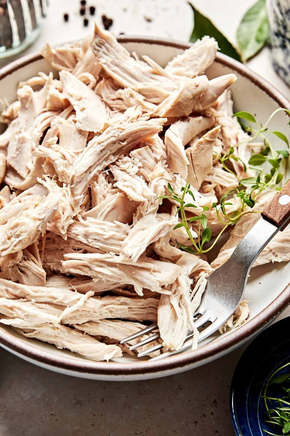 A close-up angled shot of a shallow stoneware bowl of shredded chicken. A sprig of thyme sits atop the chicken, and a small blue dish of herbs sits alongside the bowl.