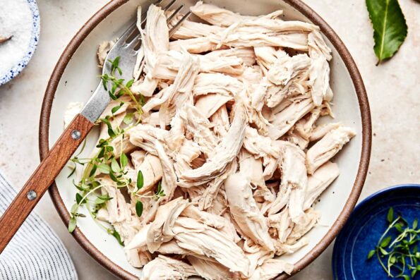 An overhead shot of shredded chicken with a fork in a shallow stoneware bowl atop an off-white surface. The bowl is surrounded by a small blue bowl of herbs, a dish of salt and a blue and white striped cloth.