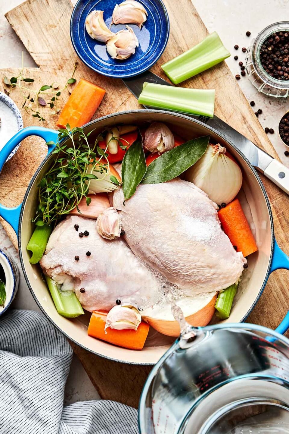 An overhead shot of ingredients to poach chicken in a large Dutch oven: raw chicken, carrots, celery, onion, peppercorns, fresh herbs, garlic and bay leaves. The Dutch oven sits atop a wooden board on an off-white surface. A glass measuring cup of water is being poured over the ingredients in the Dutch oven.