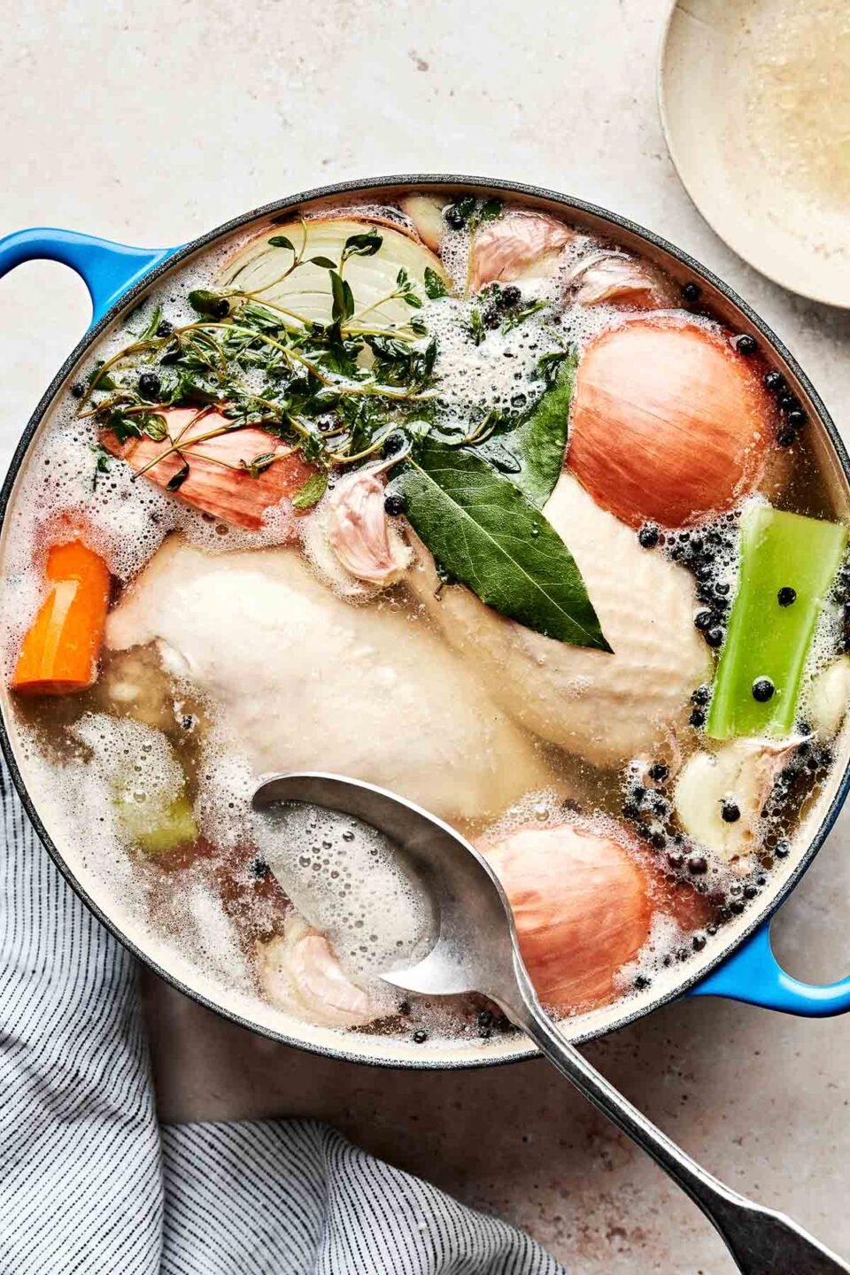 An overhead shot of poaching chicken, carrots, onions, celery, peppercorns and fresh herbs simmering in a Dutch oven atop an off-white textured surface.
