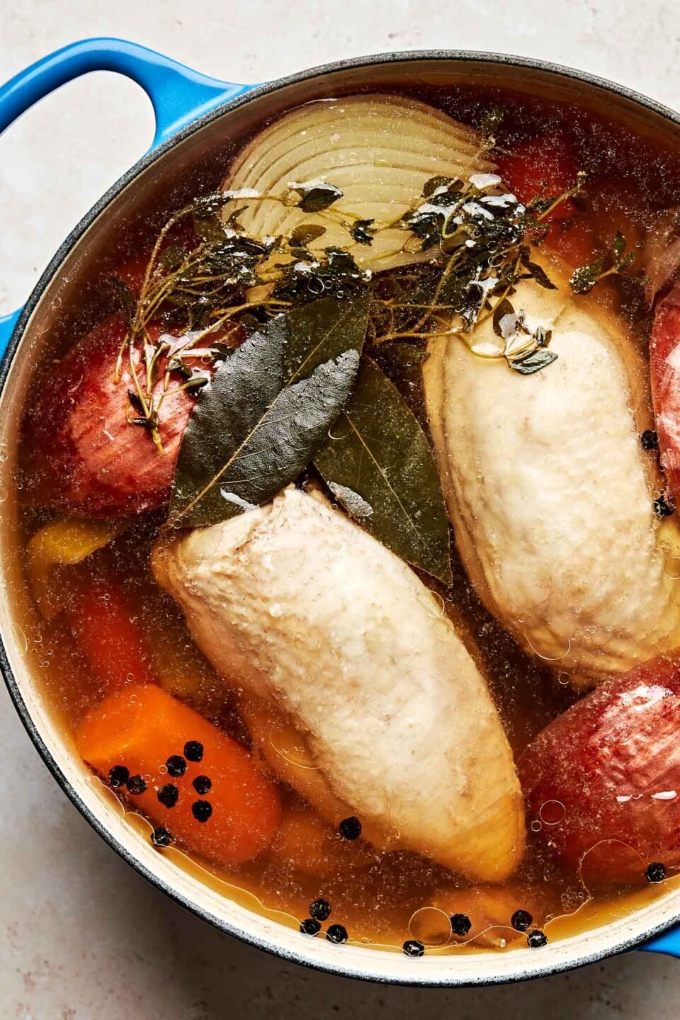 An overhead shot of poaching chicken in a broth of vegetables and herbs in a blue Dutch oven atop an off-white surface.