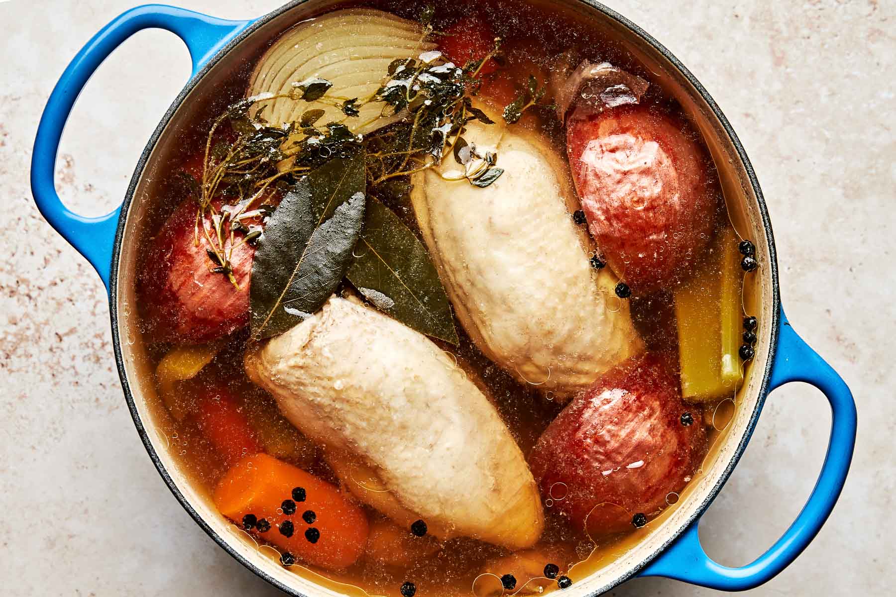 An overhead shot of poaching chicken in a broth of vegetables and herbs in a blue Dutch oven atop an off-white surface.