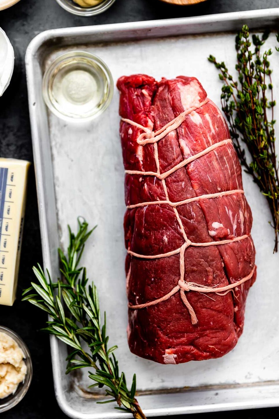 An overhead shot of ingredients displayed on a sheet pan and a dark grey surface: a beef tenderloin wrapped in twine, fresh thyme, fresh rosemary, a stick of butter, and dishes of pepper, dijon mustard, salt, sour cream, horseradish and lemon juice.