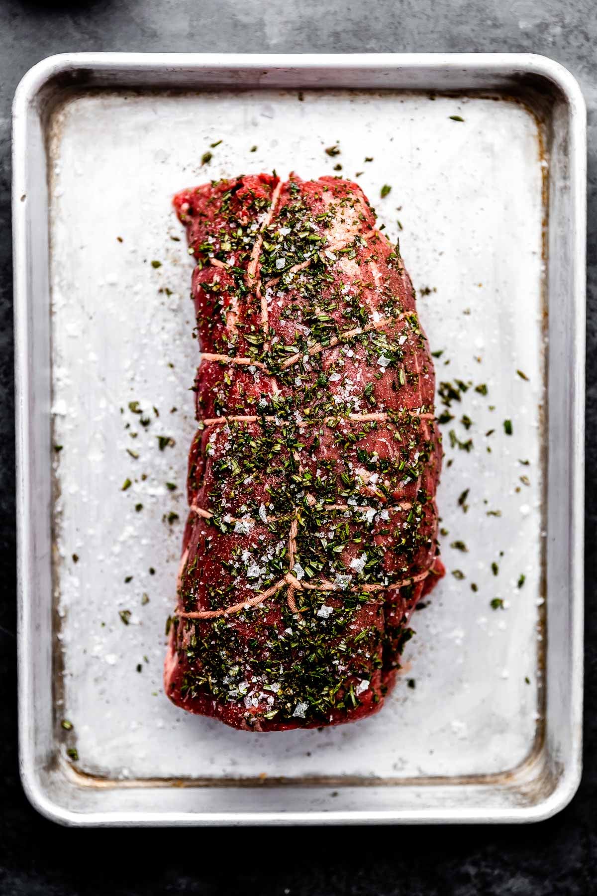 An overhead shot of a seasoned beef tenderloin wrapped with kitchen twine on a sheet pan atop a dark grey surface.