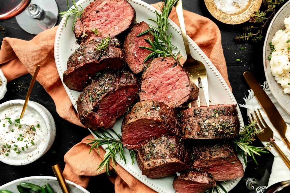 An overhead shot of large pieces of beef tenderloin on a white oval platter atop an orange cloth on a black surface. Dishes of mashed potatoes, horseradish sauce, asparagus and sea salt sit alongside the platter.