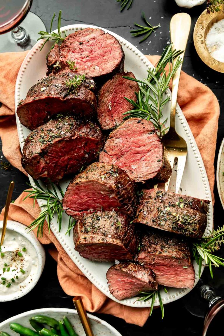 An overhead shot of large pieces of beef tenderloin on a white oval platter atop an orange cloth on a black surface. Dishes of mashed potatoes, horseradish sauce, green beans and sea salt sit alongside the platter.