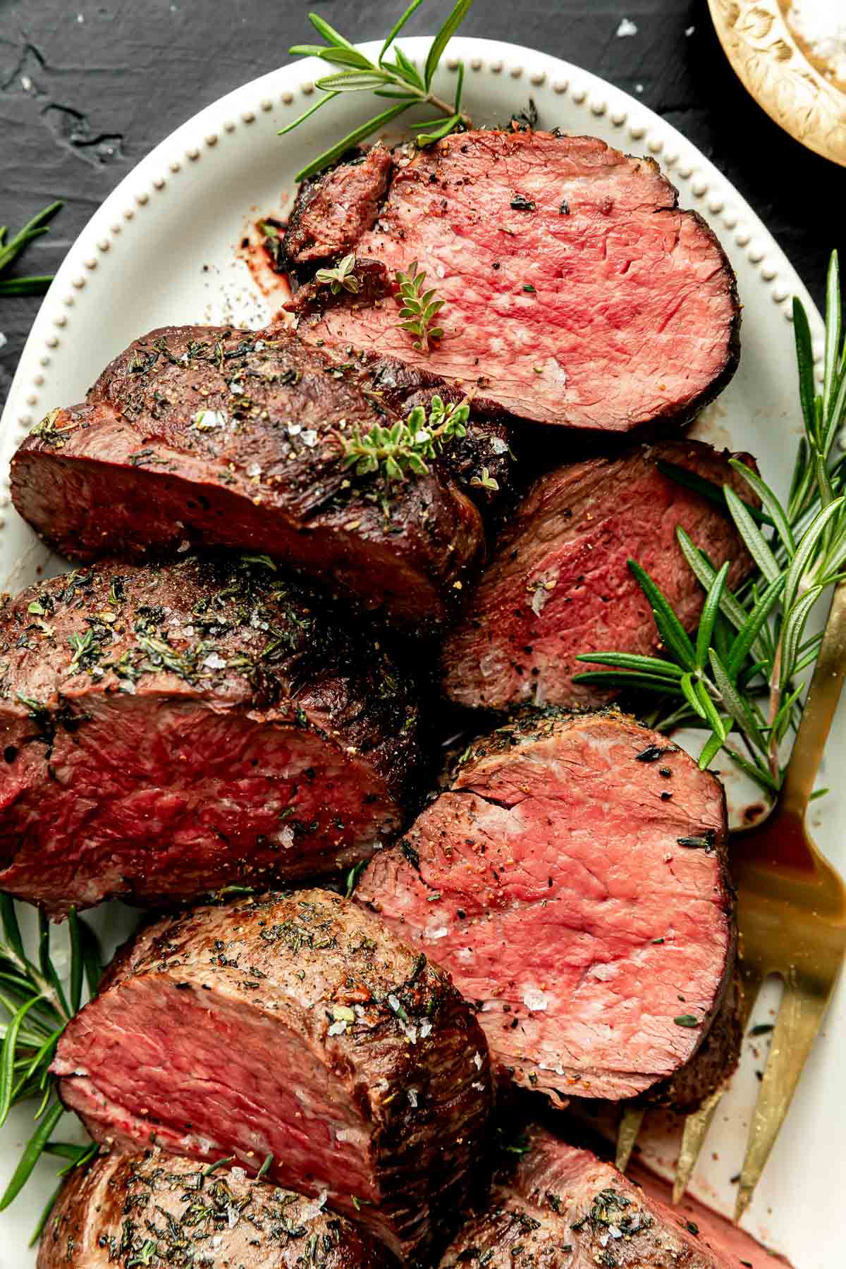 An overhead shot of large pieces of beef tenderloin on a white oval platter on a black surface.