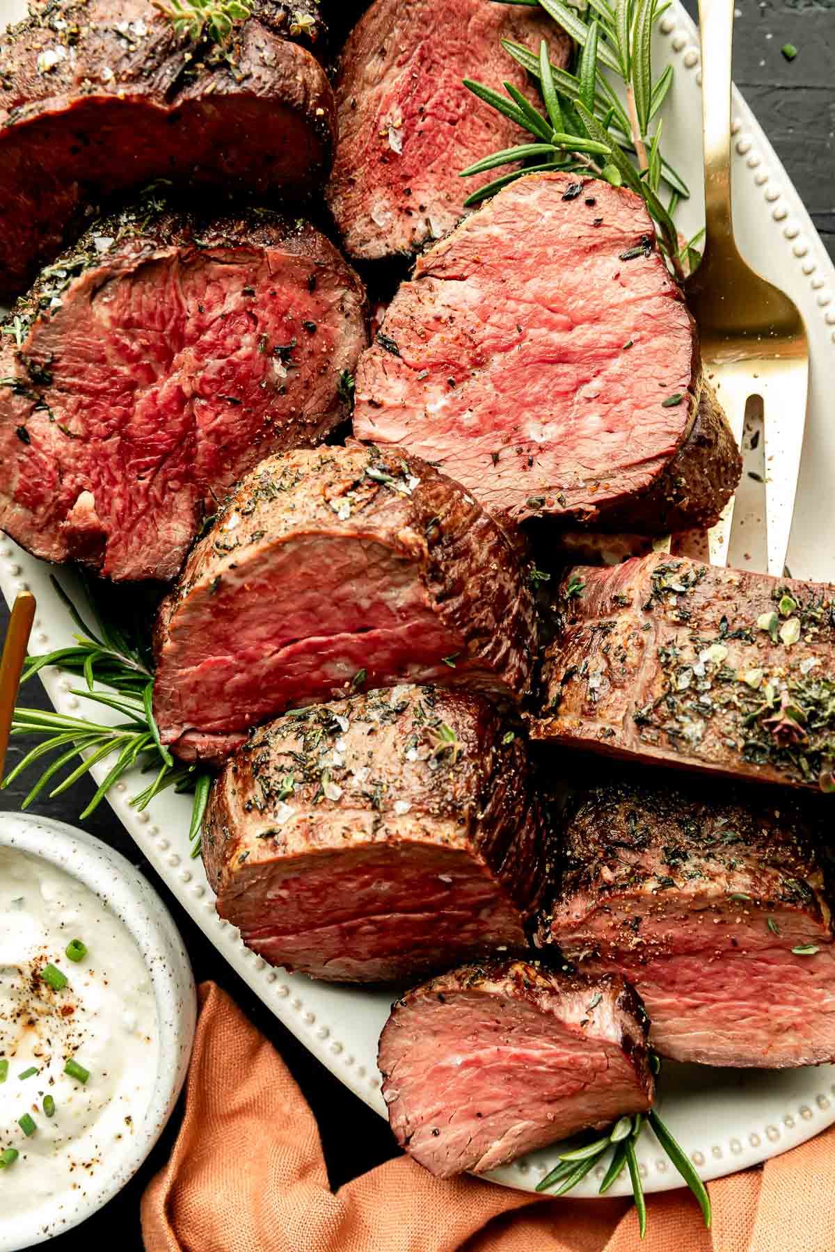 An overhead shot of large pieces of beef tenderloin on a white oval platter atop an orange cloth on a black surface. A small bowl of sauce sits alongside the platter.