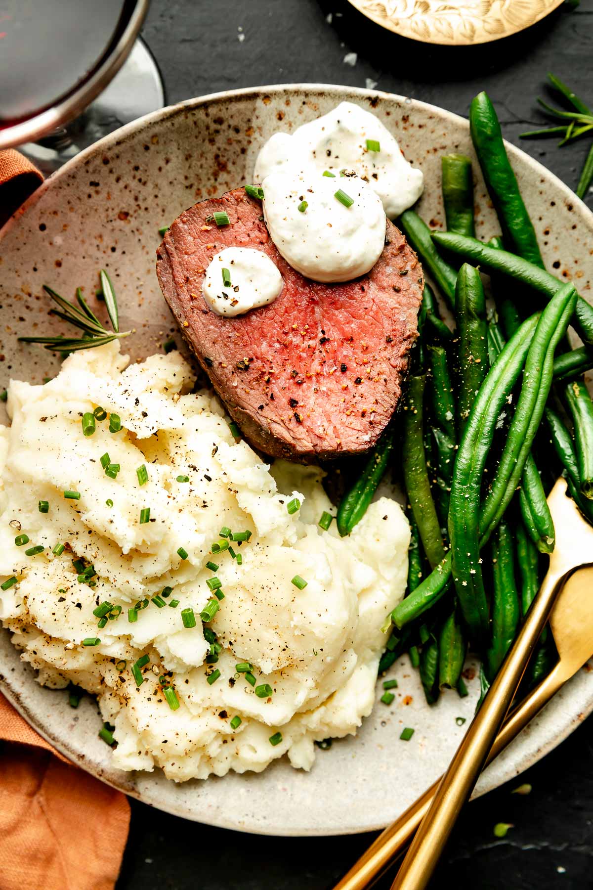 An overhead shot of a slice of beef tenderloin with sauce, cooked green beans, and mashed potatoes on a stoneware plate atop a black surface.