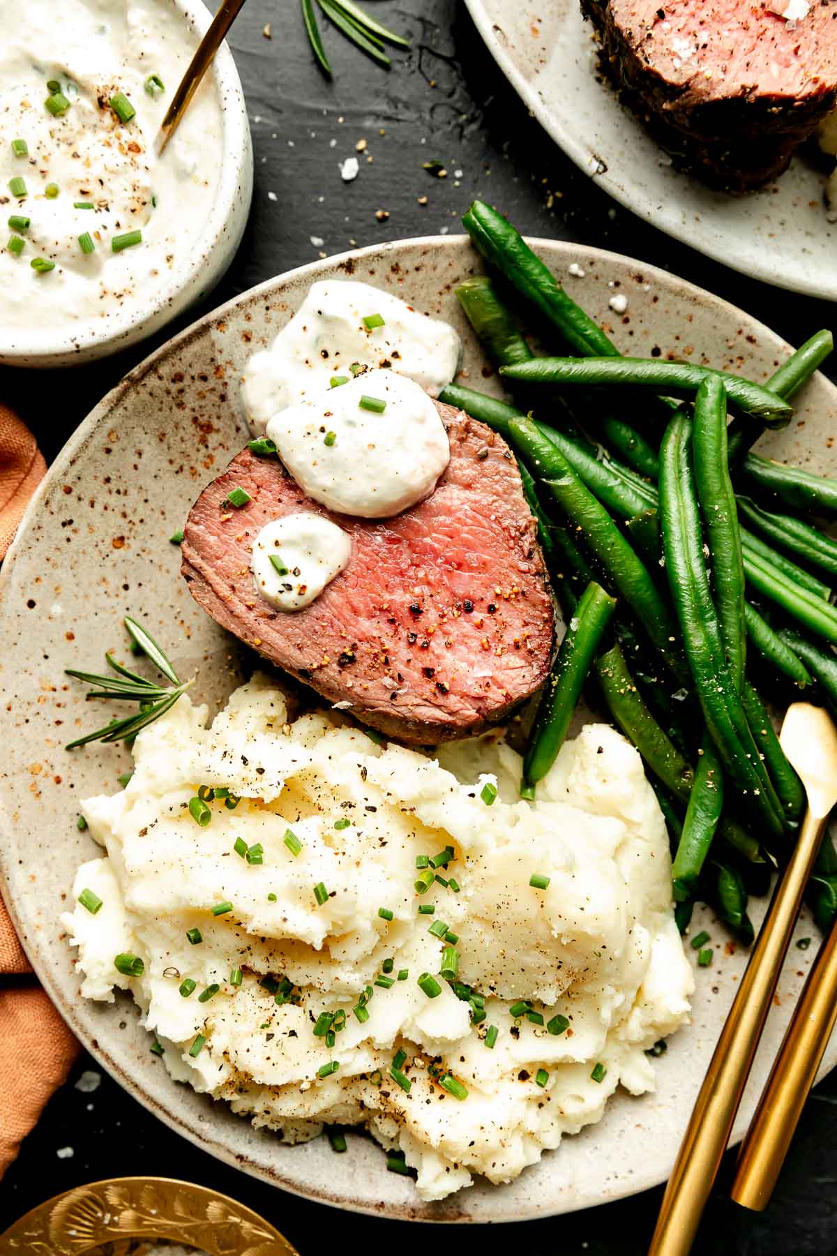 An overhead shot of a slice of beef tenderloin with sauce, cooked green beans, and mashed potatoes on a stoneware plate atop a black surface. A bowl of sauce and the platter of tenderloin sit beside the plate.