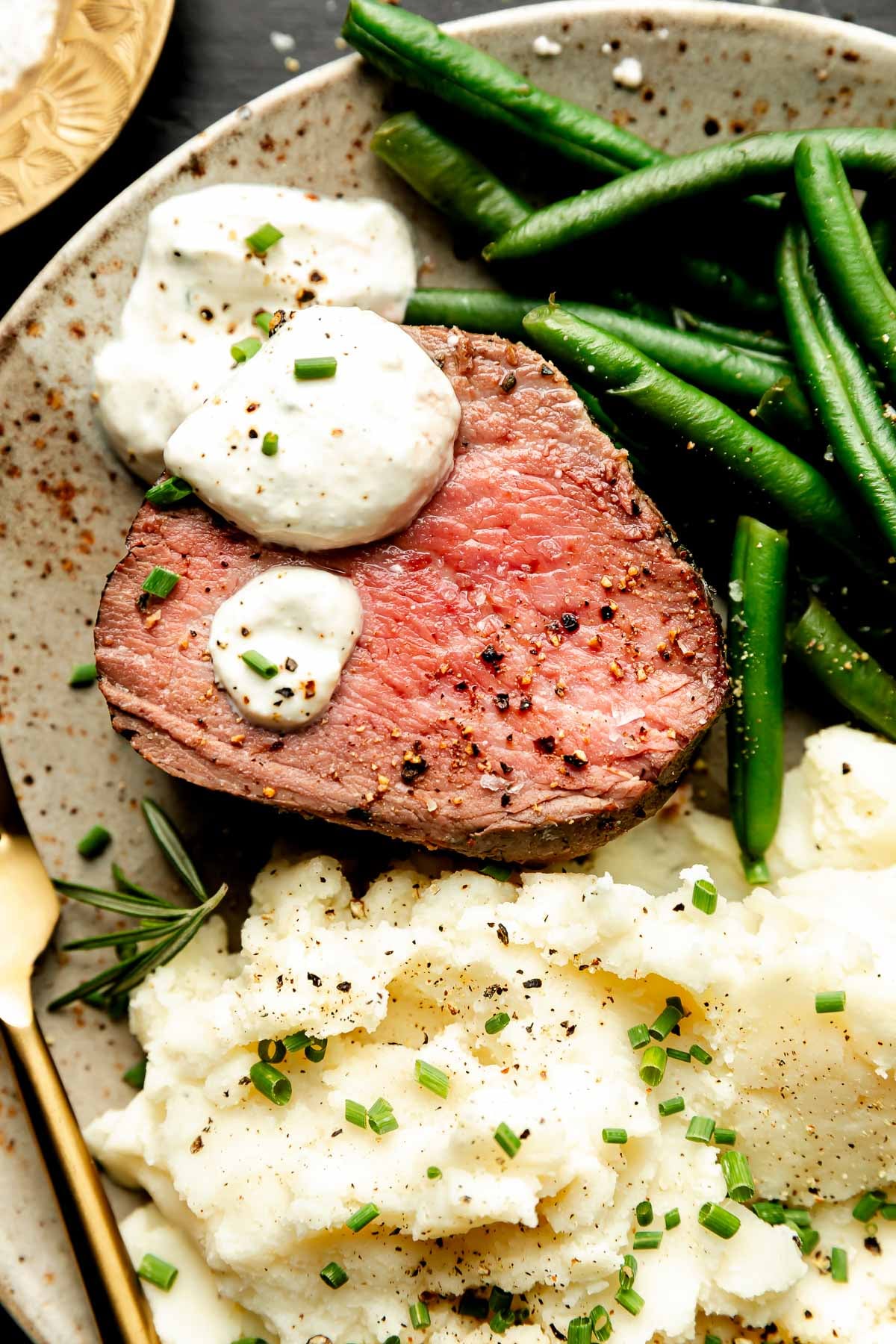 A close-up overhead shot of a slice of beef tenderloin topped with sauce, mashed potatoes and green beans on a stoneware plate.