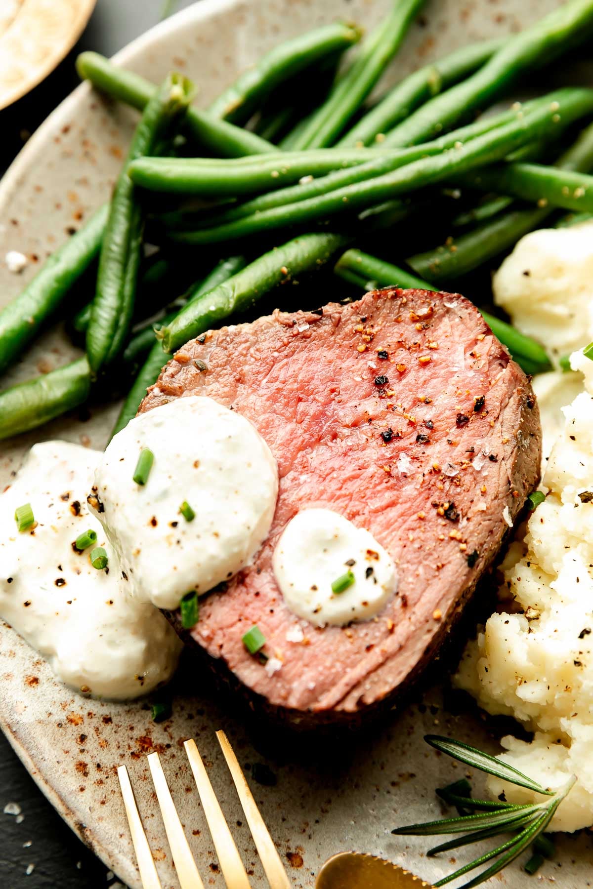 A close-up shot of a slice of beef tenderloin topped with sauce, mashed potatoes and green beans on a stoneware plate.