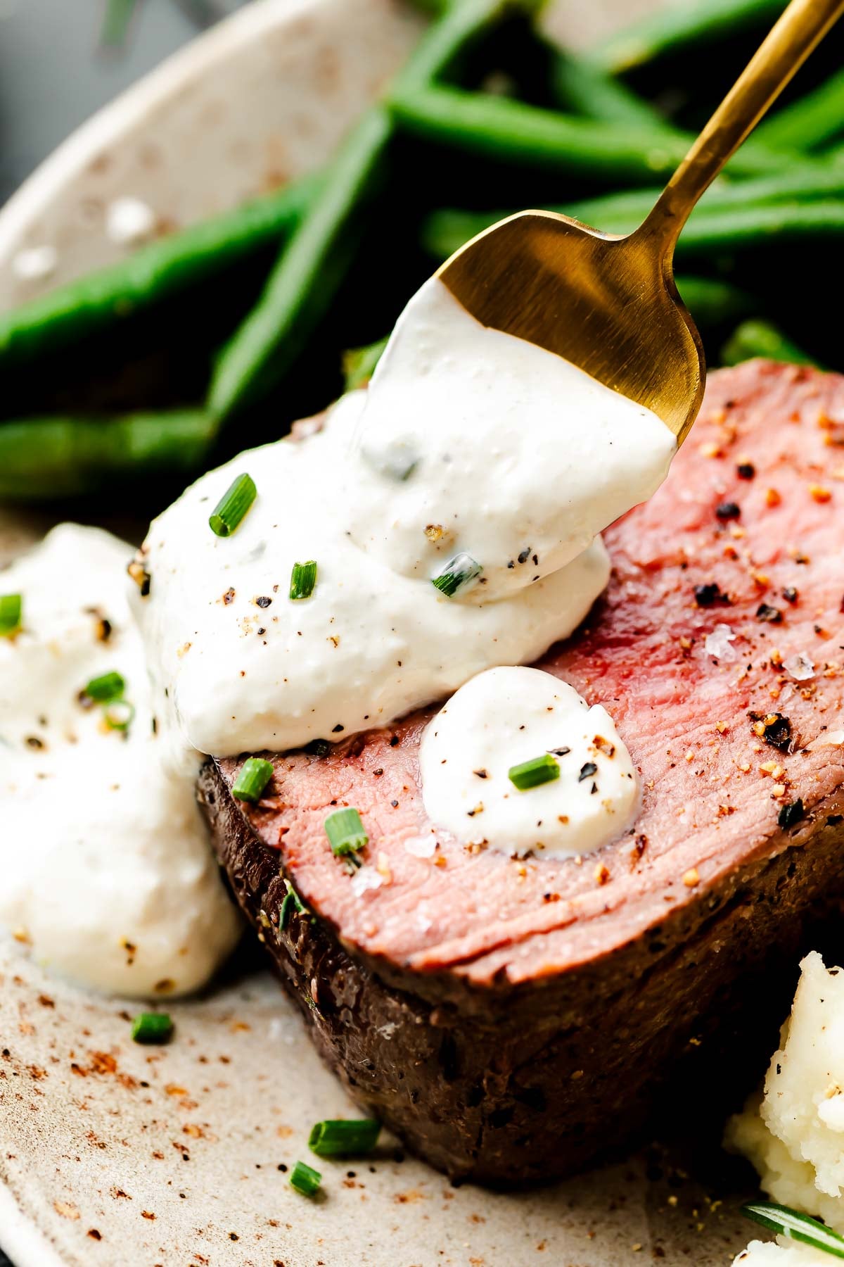 A close-up macro shot of a slice of beef tenderloin being topped with sauce from a gold spoon alongside mashed potatoes and green beans on a stoneware plate.