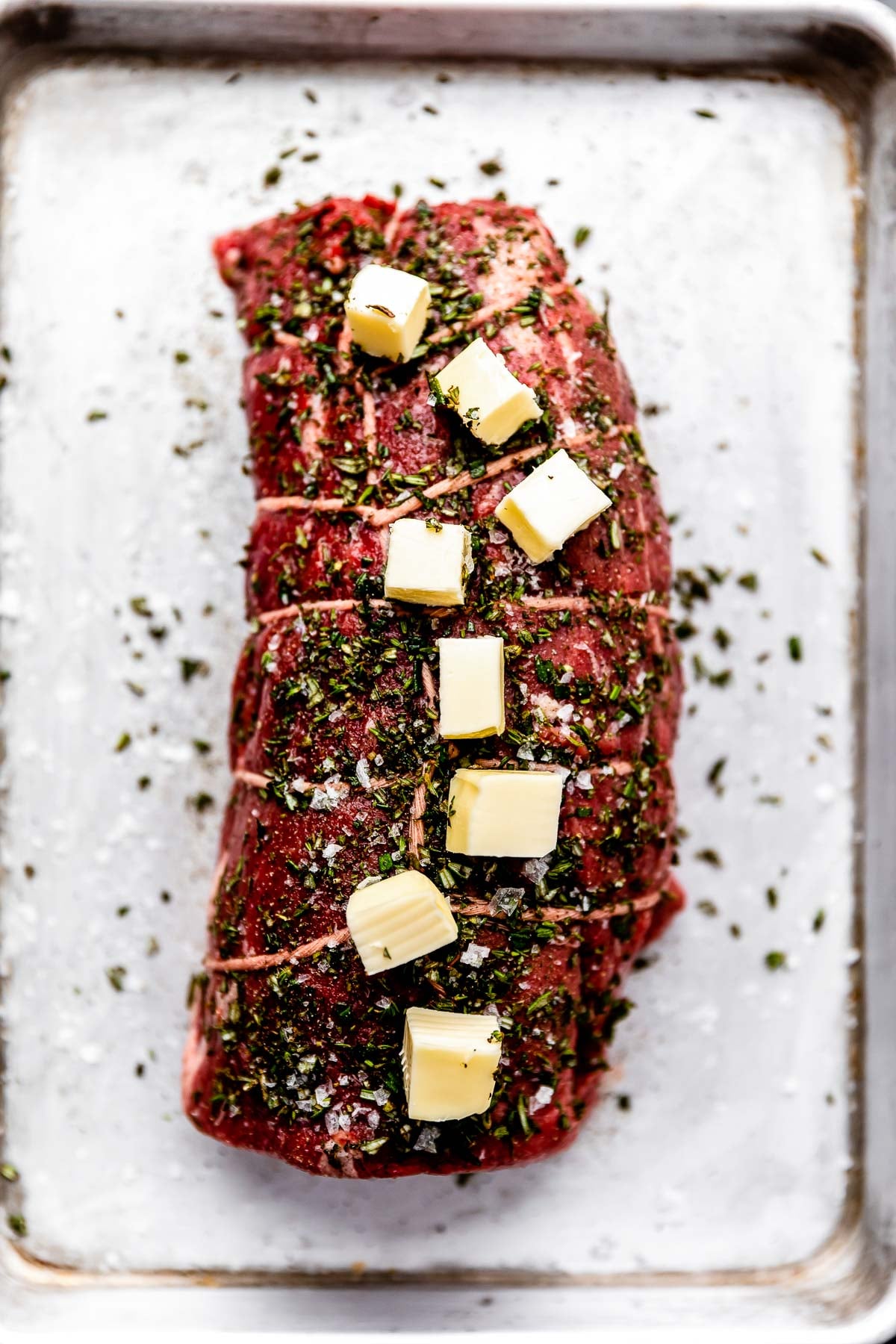 An overhead shot of a seasoned beef tenderloin wrapped in twine and topped with cubes of butter on a small sheet pan.