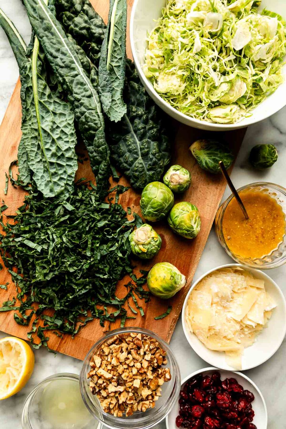 An overhead shot of ingredients displayed on a wooden cutting board and a white marbled surface: whole kale leaves and chopped kale, whole and shredded brussels sprouts, and dishes of parmesan, dressing, walnuts, dried cranberries and lemon juice.