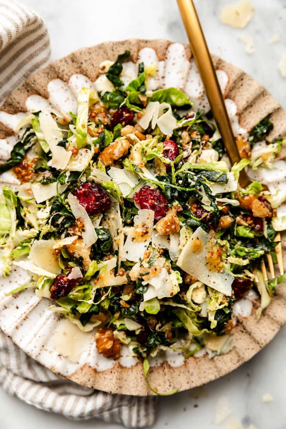An overhead shot of a serving of kale and brussels sprouts salad topped with parmesan and black pepper on a scalloped brown and white plate atop a white surface. A striped cloth sits alongside the plate.