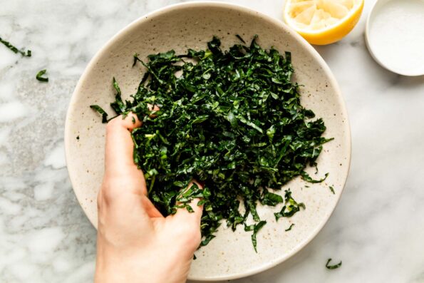An overhead shot of a woman's hand massaging kale with lemon juice in a large stoneware bowl atop a white marbled surface.