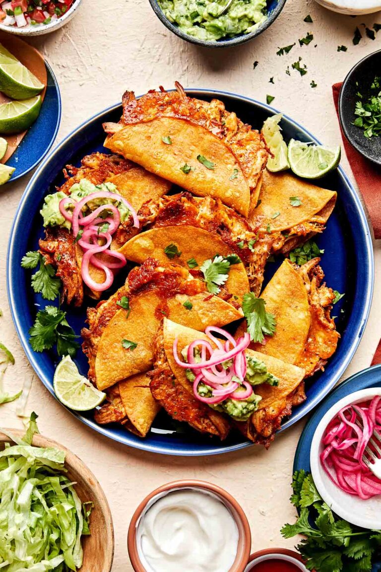 A blue plate filled with baked crispy chicken tacos topped with pickled onions, jalapeños, and cilantro, garnished with lime wedges. Surrounding the plate are bowls of lettuce, guacamole, sour cream, and salsa.