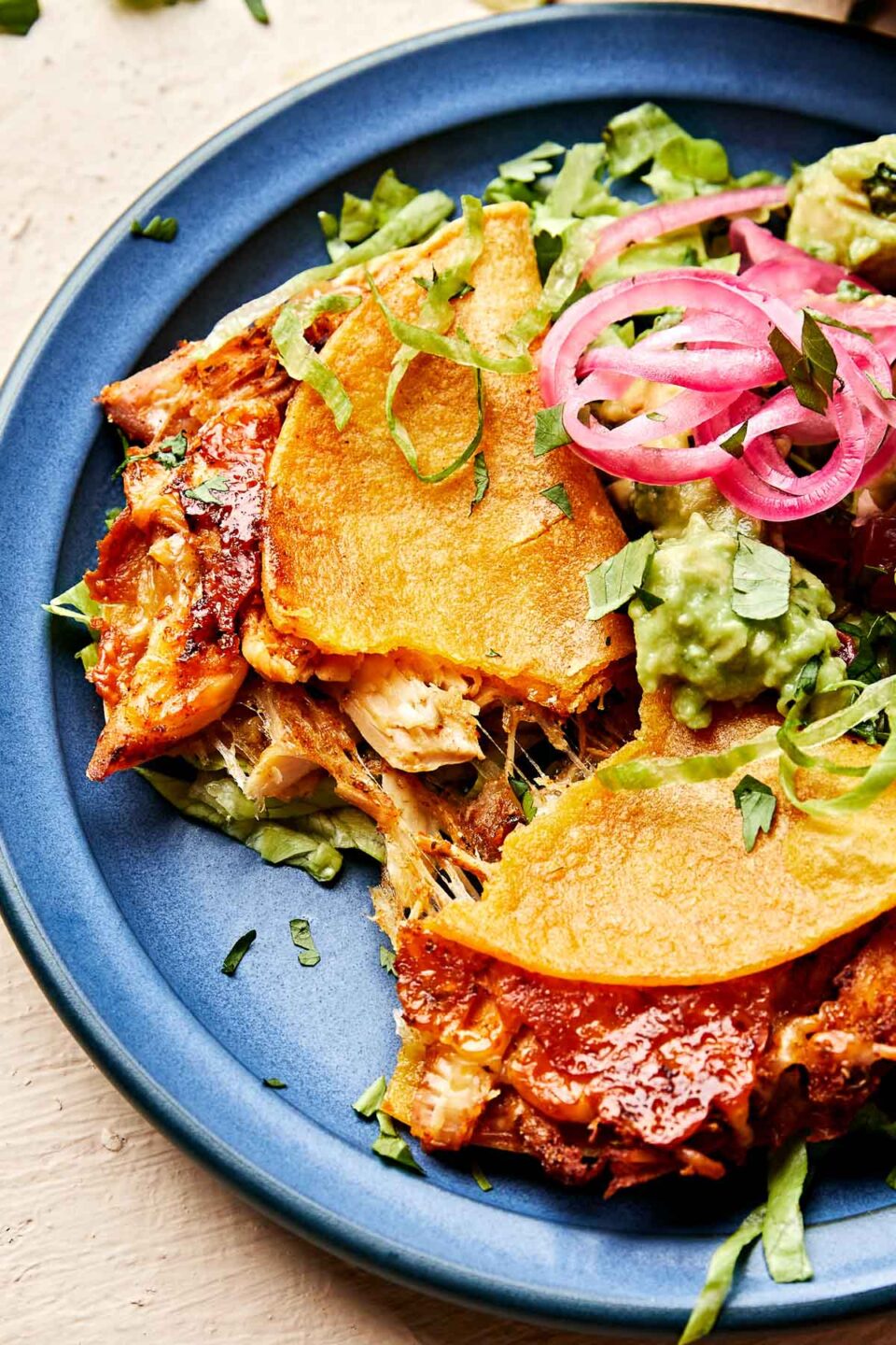 A close-up shot of two crispy baked chicken tacos topped with guacamole, lettuce and pickled red onions on a blue plate. 