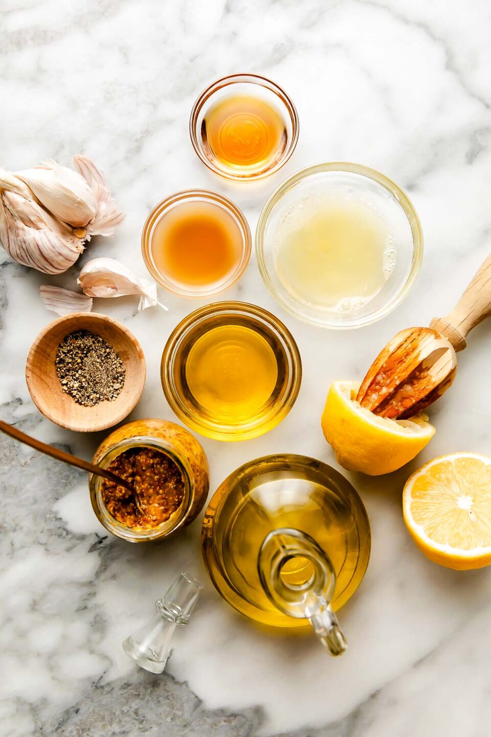 An overhead shot of ingredients displayed in small bowls and dishes on a white marbled surface: olive oil, whole grain mustard, apple cider vinegar, lemon juice, a juiced lemon, garlic, black pepper and maple syrup.