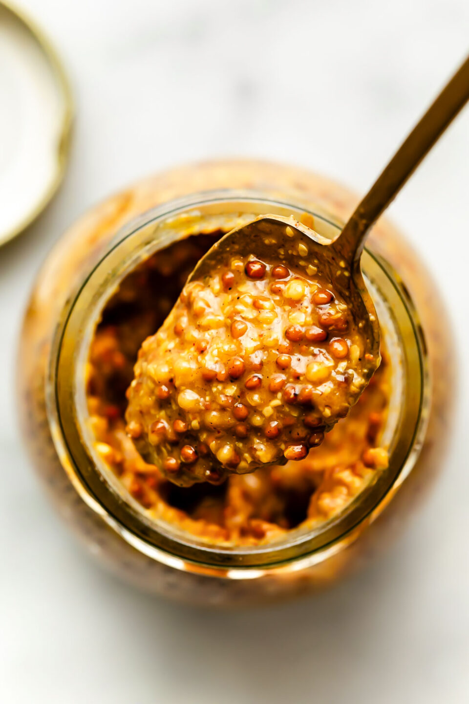 An overhead macro shot of a spoon full of whole grain mustard over a jar on a white marbled surface.