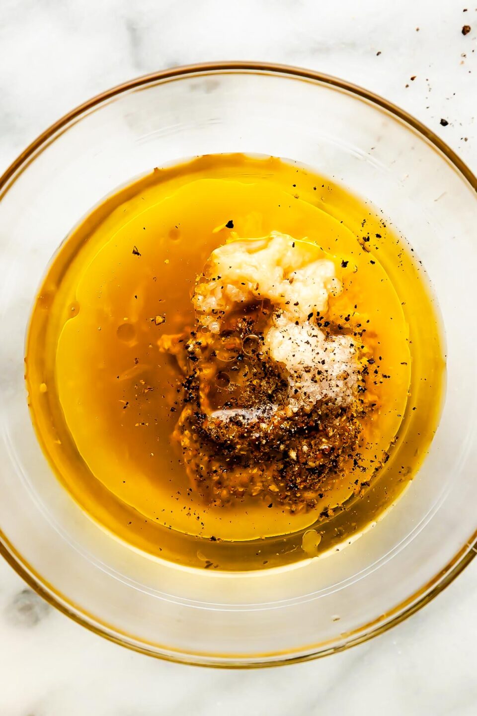 An overhead shot of ingredients in a small glass bowl: whole grain mustard, lemon juice, apple cider vinegar, maple syrup, black pepper, and garlic.