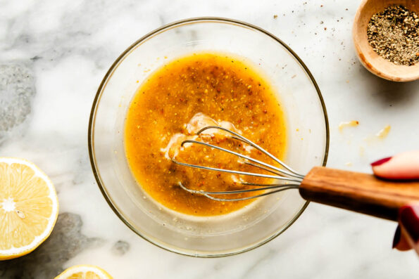 An overhead shot of vinaigrette being whisked in a glass bowl atop a white marbled surface. A halved lemon and a small bowl of pepper sit alongside the bowl.