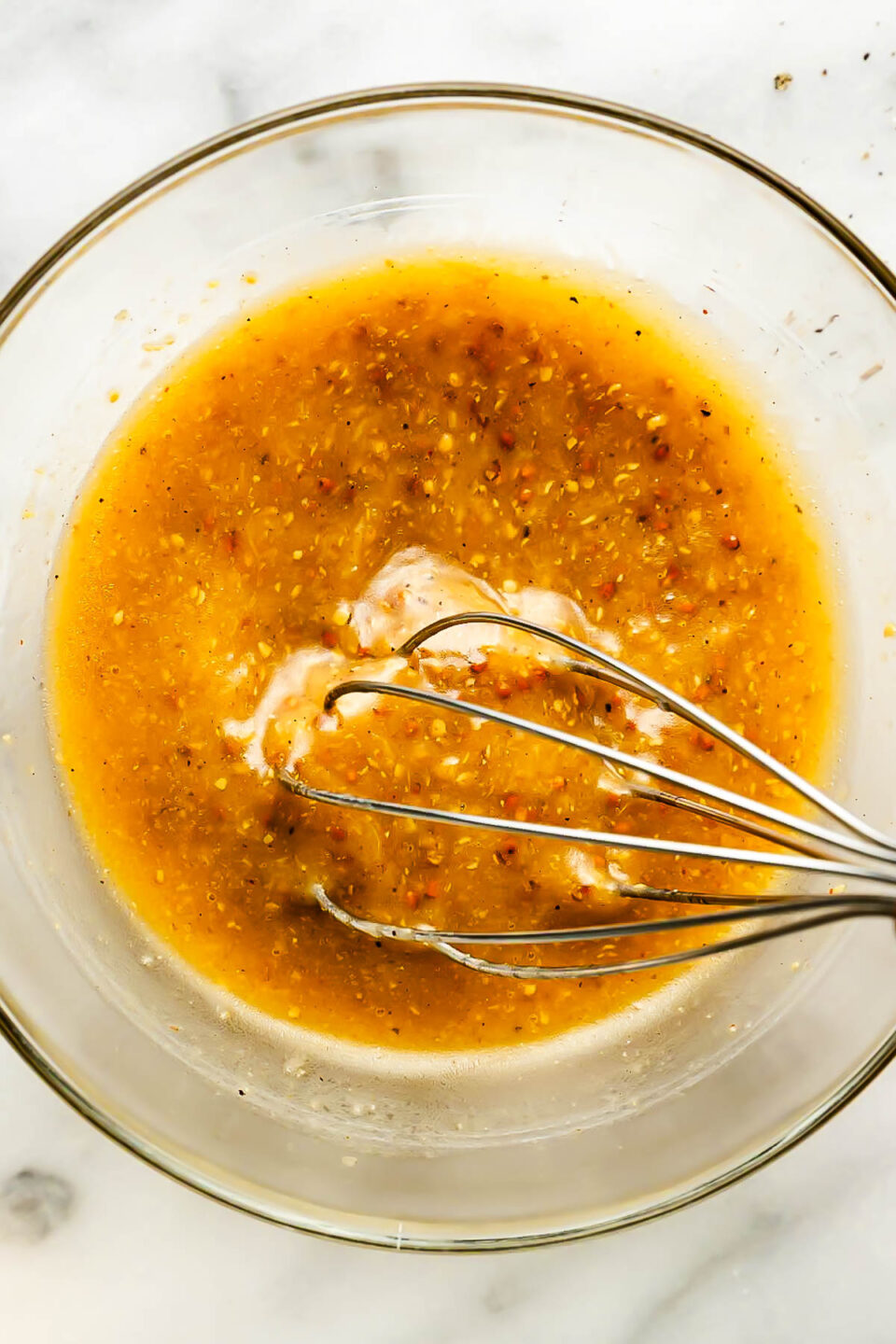 An overhead shot of vinaigrette being whisked in a glass bowl atop a white marbled surface.