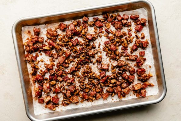 An overhead shot of roasted candied pancetta spiced nuts on a small metal sheet pan atop an off-white textured surface.