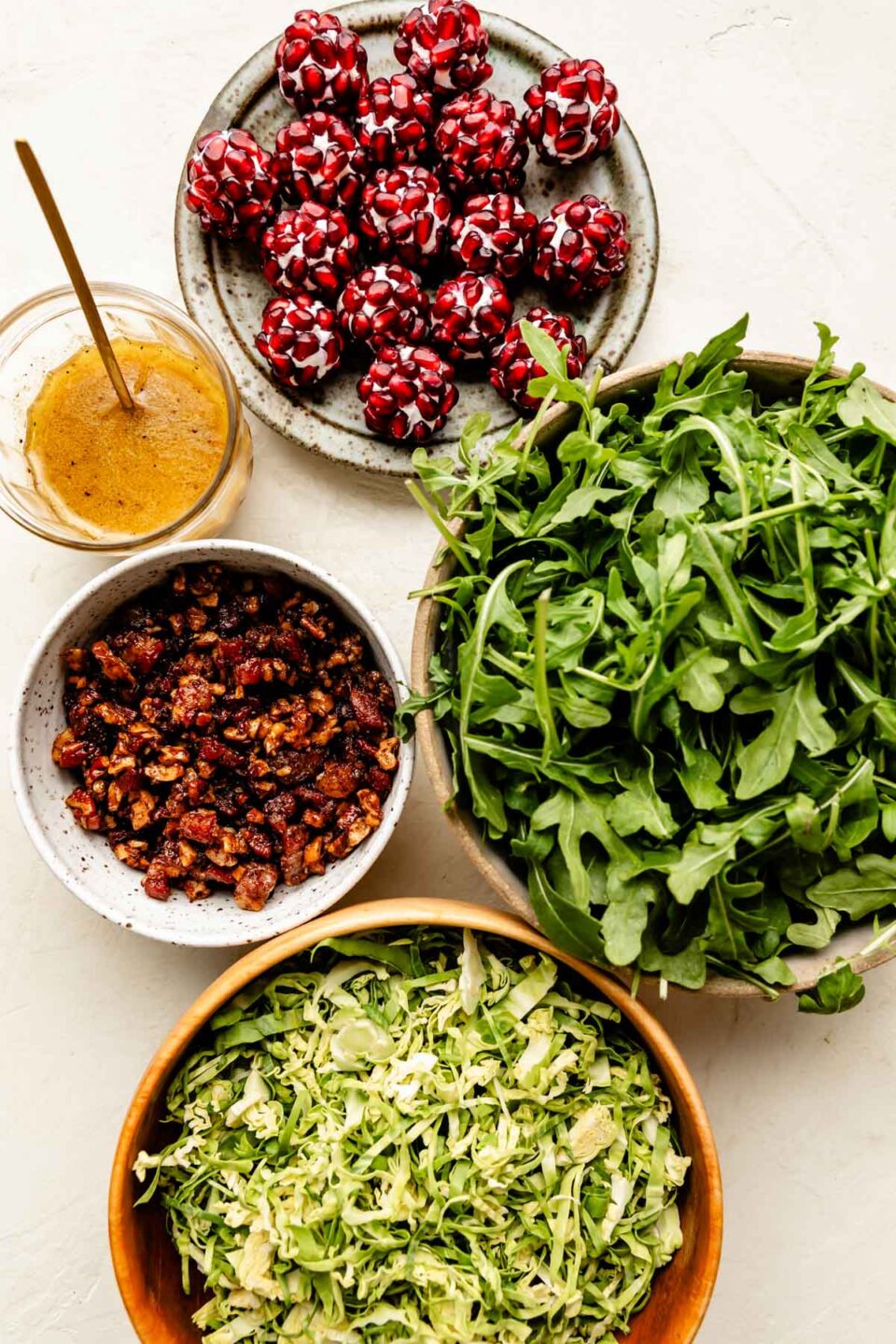 An overhead shot of ingredients displayed atop an off-white surface: baby arugula, shaved brussels sprouts, candied nuts, a jar of dressing and a plate of pomegranate goat cheese balls.