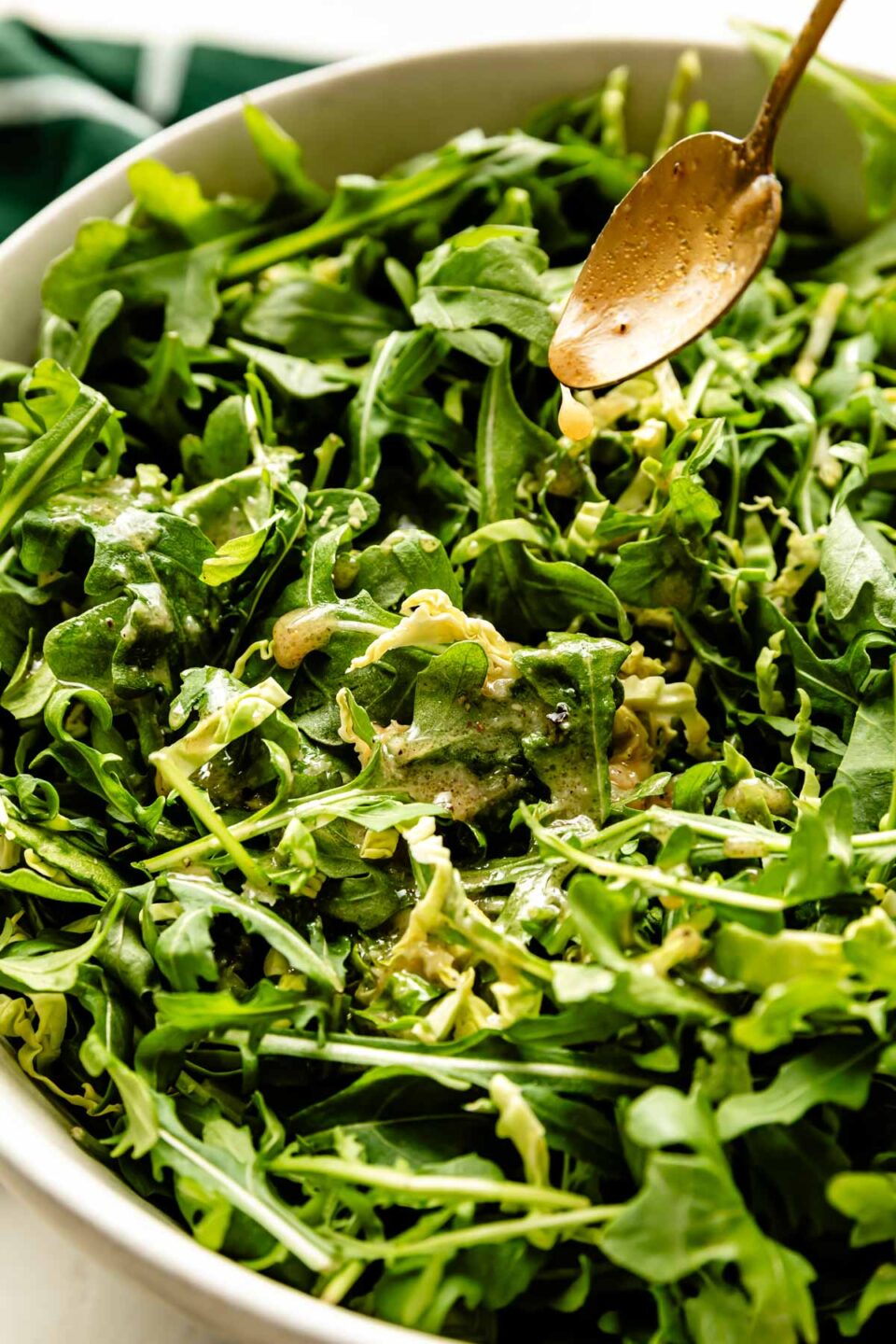 An overhead shot of dressing being drizzled from a gold spoon over a large bowl of arugula.