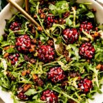 An overhead shot of an assembled Christmas salad: arugula, candied nuts, shaved brussels sprouts, and pomegranate goat cheese balls. The salad is in a large white bowl atop a green plaid cloth on an off-white surface.