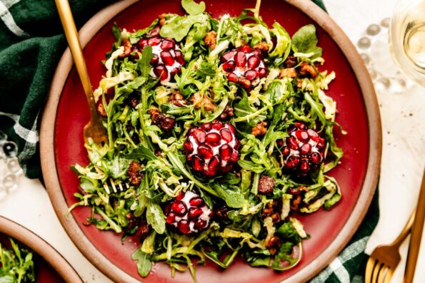 An overhead shot of a serving of Christmas salad on a dark red plate atop a green plaid cloth on an off-white surface.