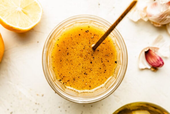 An overhead shot of a glass jar of maple dijon vinaigrette atop an off-white surface. A juiced lemon and garlic cloves sit alongside the jar.