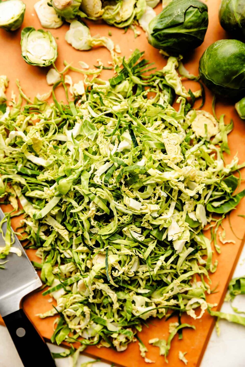 An overhead shot of shredded Brussels sprouts and whole Brussels sprouts with a chef's knife on a light brown cutting board atop an off-white surface.
