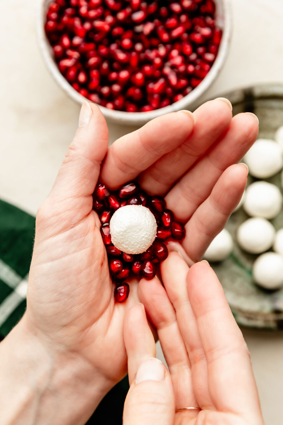 An overhead shot of a woman's hand holding a ball of goat cheese and pomegranate arils, forming a pomegranate goat cheese ornament. A plate of goat cheese balls and a bowl of pomegranate arils sits in the background.