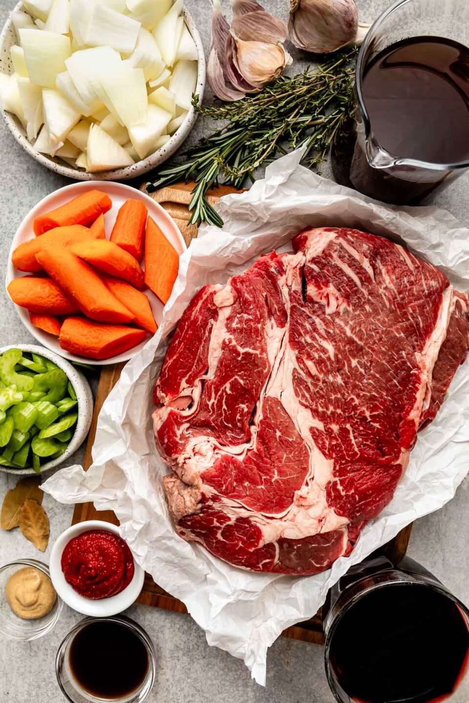 An overhead shot of ingredients displayed on a light grey textured surface: beef on butcher paper, carrots, celery, onion, fresh herbs, garlic, bay leaves, red wine, beef stock, worcestershire sauce, and dijon mustard.