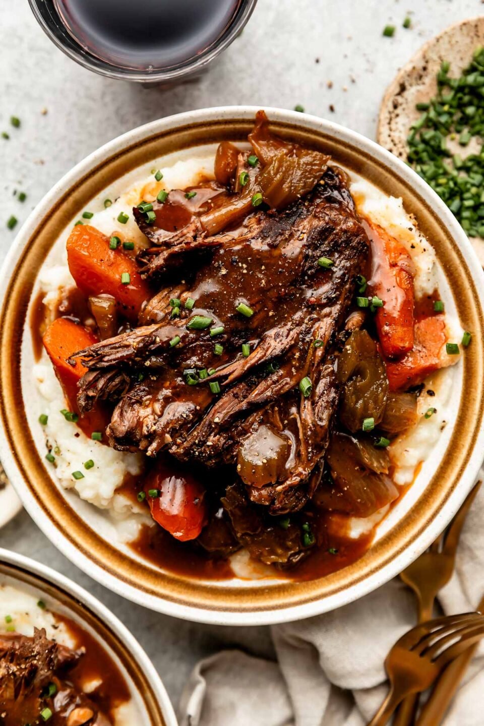An overhead shot of a serving of braised beef topped with braising liquid on a bed of potatoes and cooked vegetables on a stoneware plate. The plate sits alongside a dish of chives, a dish of pepper and gold silverware on a light grey surface.
