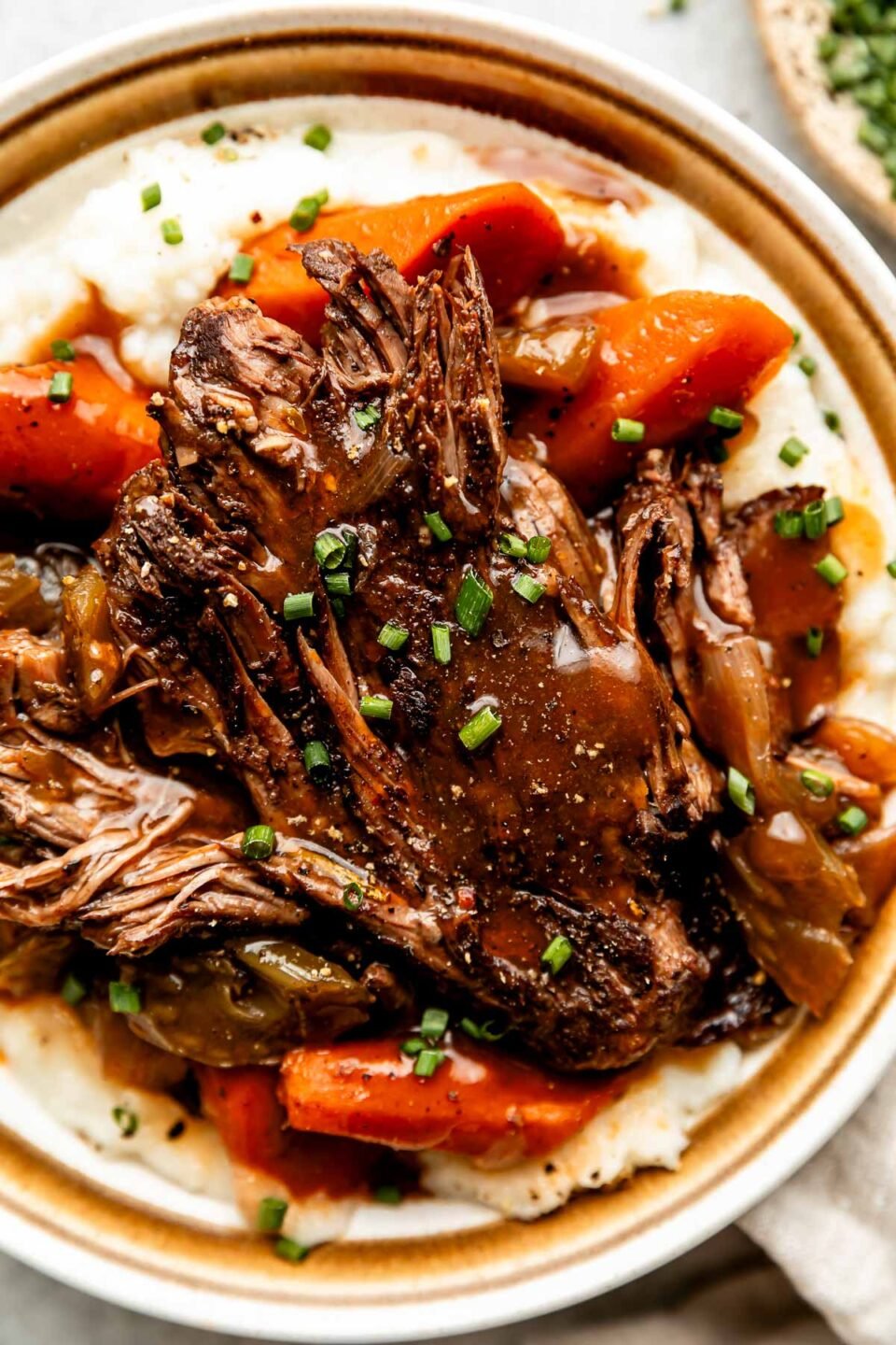 An up-close overhead shot of a serving of braised beef topped with braising liquid on a bed of potatoes and cooked vegetables on a stoneware plate.