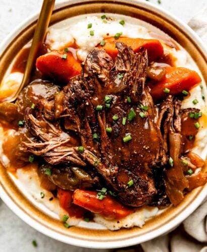 An overhead shot of a serving of braised beef topped with braising liquid on a bed of potatoes and cooked vegetables on a stoneware plate. The plate sits alongside a beige cloth on a light grey surface.