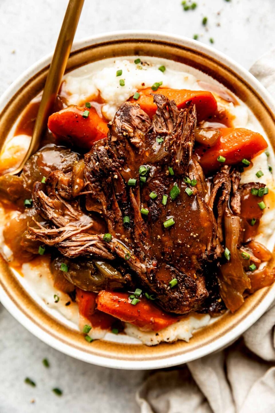 An overhead shot of a serving of braised beef topped with braising liquid on a bed of potatoes and cooked vegetables on a stoneware plate. The plate sits alongside a beige cloth on a light grey surface.