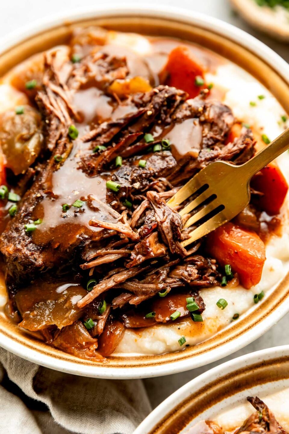 A close up side shot of a fork grabbing a bite of braised beef from a plate of beef on a bed of mashed potatoes and vegetables.
