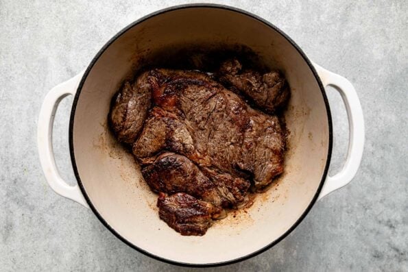 An overhead shot of seared beef in a Dutch oven atop a light grey textured surface.